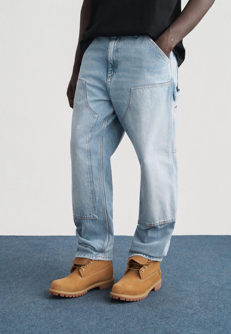Homme portant un jean de travail bleu clair avec des genoux renforcés et des bottes à lacets couleur cuir clair, debout sur un tapis bleu, une main dans la poche.