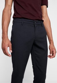 Pantalon bleu marine foncé présentant une texture lisse, une coupe ajustée, des poches avant, une fermeture à bouton unique et des passants pour ceinture.