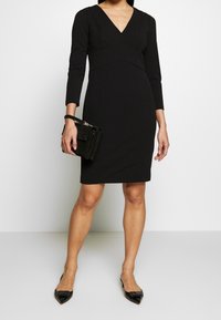 Musta vartalonmyötäinen mekko pitkillä hihoilla ja V-pääntiellä, jossa on istuva vyötärö ja sileä pinta. Yhdistetty pieni musta käsilaukku.