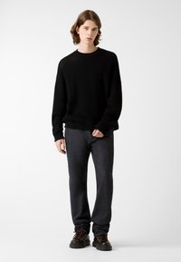 Schwarzer, strukturierter Pullover mit Rundhalsausschnitt, kombiniert mit dunklen Jeans aus Denim. Das Modell trägt robuste schwarze Schnürstiefel. Minimalistischer Hintergrund.