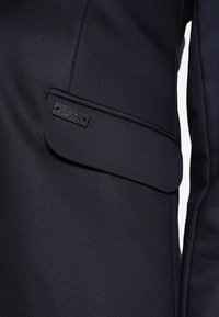 Marinblå blazer med en klaffficka med en sydd kant, prydd med en svart emblemplatta. Slät tyg med subtil lyster.