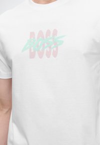 Λευκή μπλούζα με επικαλυπτόμενο κείμενο "BOSS" σε ροζ και ανοιχτό μπλε στο στήθος, φορεμένη από άτομο που εμφανίζεται από το λαιμό μέχρι τη μέση.