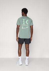 Zaļš kokvilnas T-krekls ar drukātu grafiku uz muguras, kurā attēlots "EK" logo, teksts "NEKADS cilvēks nav ierobežots" un Nike zoss. Kombinēts ar melnām šortiem.
