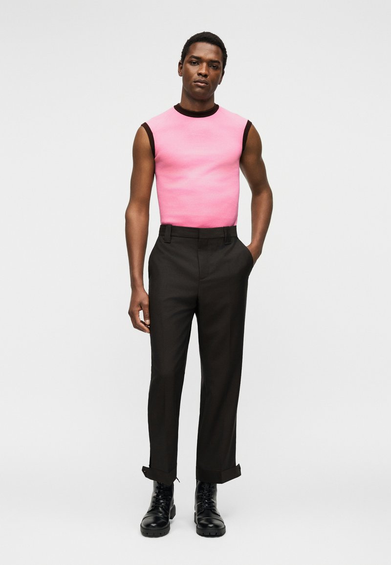 Man wearing sleeveless pink top with black trim, black high-waist pants with cuffs, and black lace-up boots, standing with one hand in pocket.