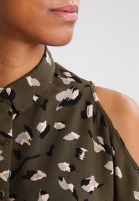 Gros plan sur une personne portant un haut sans manches vert olive à épaules dénudées avec un motif abstrait noir, blanc et beige et un col à boutons.