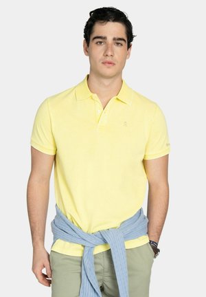 Joven con polo amarillo, suéter azul claro atado a la cintura y pantalones verde claro, de pie contra un fondo liso.