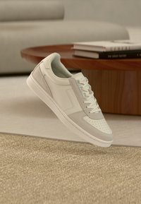White and light gray athletic sneakers with a smooth leather upper, perforated toe, padded collar, and flat laces, displayed on a textured surface.
