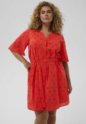 Kobieta o kręconych włosach, ubrana w czerwone, sięgające kolan sukienkę na guziki z krótkimi rękawami i paskiem wiązanym w talii, stoi z jedną ręką na biodrze.