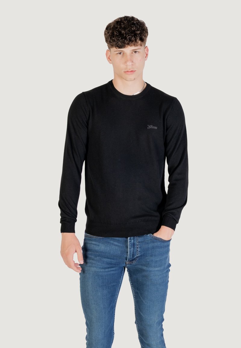 Maglione nero lavorato a maglia con maniche lunghe e scollatura tonda, con polsini e orlo a costine, indossato con jeans blu. Piccolo logo sul petto.