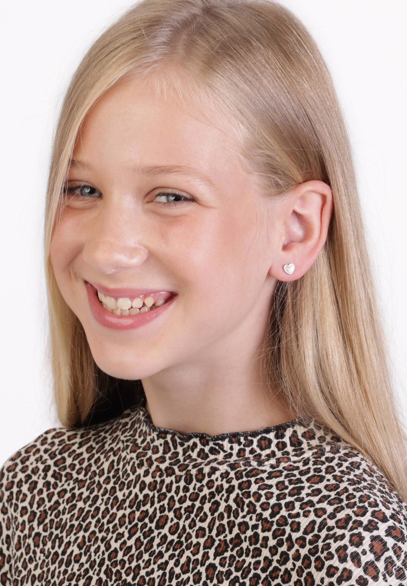 Sterling zilveren hartvormige oorbellen, klein en glanzend, gedragen door een meisje met lang, steil blond haar, gekleed in een top met leopardprint.