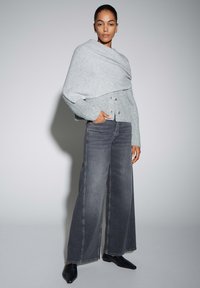 Pull en maille gris clair avec un design drapé et deux boutons ; associé à un jean denim gris foncé à jambes larges et des chaussures noires à bouts pointus.