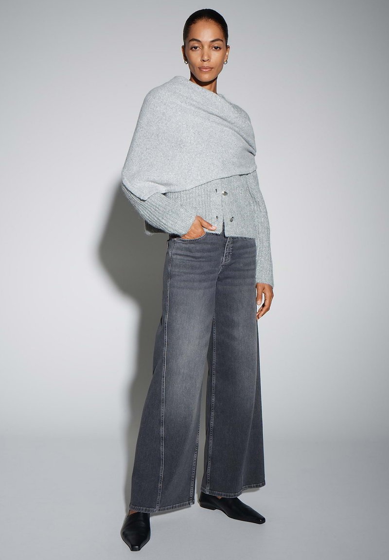 Pull en maille gris clair avec un design drapé et deux boutons ; associé à un jean denim gris foncé à jambes larges et des chaussures noires à bouts pointus.