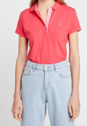 Vrouw draagt een koraalroze poloshirt met korte mouwen, ingestopt in een lichtblauwe high-waisted jeans tegen een effen achtergrond.
