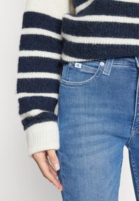 Suéter de punto a rayas en azul marino y crema con un acabado texturizado, combinado con jeans de mezclilla azul claro que cuentan con una etiqueta de logo en el bolsillo.