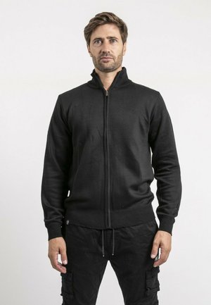 Homme aux cheveux bruns courts portant une veste noire zippée et un pantalon cargo noir, debout devant un fond blanc uni.