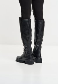 Stivali in pelle neri al ginocchio con zip laterali indossati sopra leggings neri, in piedi su una superficie bianca.