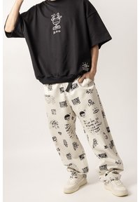 Czarna, oversize koszulka z logo; kremowe spodnie z czarnymi wzorami graficznymi; białe sneakersy; luźny krój z widocznymi kieszeniami i mankietami.