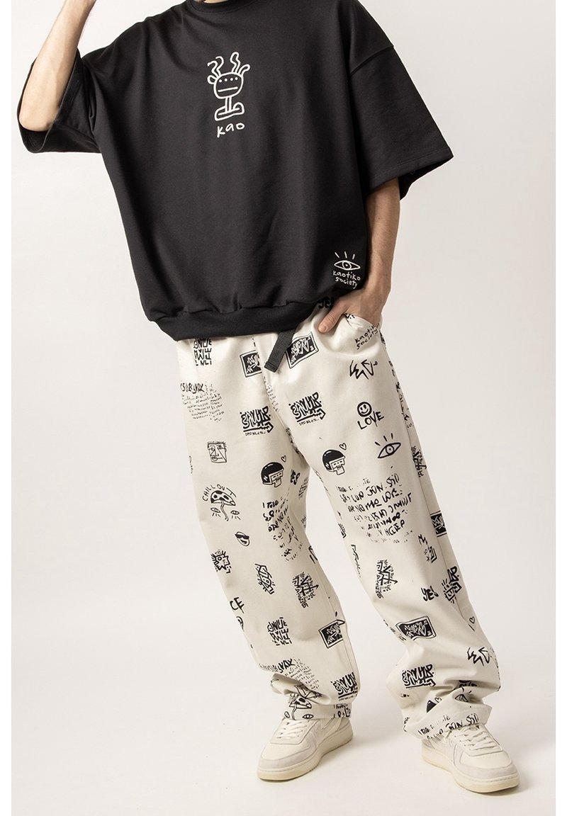 Czarna, oversize koszulka z logo; kremowe spodnie z czarnymi wzorami graficznymi; białe sneakersy; luźny krój z widocznymi kieszeniami i mankietami.