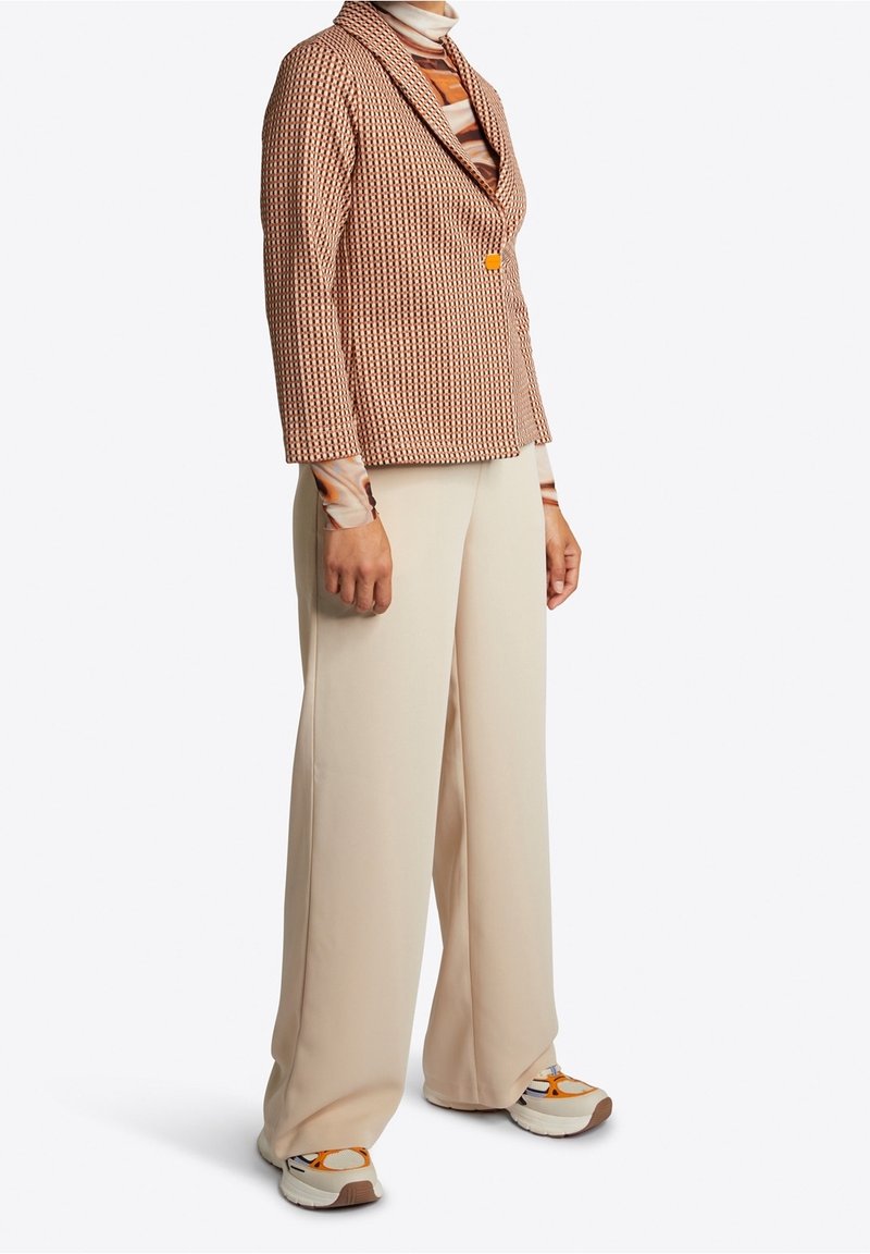 Personne portant un blazer marron texturé, un pantalon large couleur crème, un col roulé rayé coloré et des baskets blanches avec des détails orange, debout immobile.