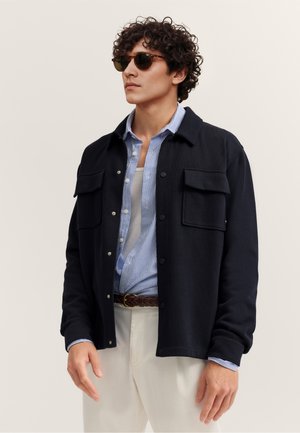 Mann mit lockigem Haar und Sonnenbrille, der eine schwarze Jacke mit Knöpfen über einem blau gestreiften Hemd und weißen Hosen vor einem einfarbigen Hintergrund trägt.