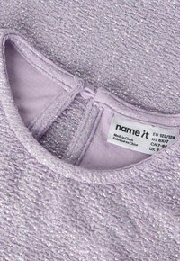 Nærbilde av lavendelfarget stoff med sølvglittertråder og en knapphempe nær kragen, som viser et klesmerke med størrelse og merkeinformasjon.