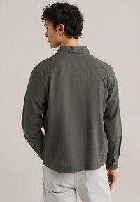 Grijze langwerpige blouse met een omgeslagen kraag, knoopmanchetten en een gestructureerde afwerking, weergegeven vanuit de achterkant.