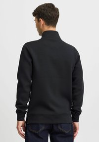 Sweat-shirt noir avec un col montant, des manches longues et une texture lisse. Présente un ourlet droit et des fentes sur les côtés pour faciliter les mouvements.