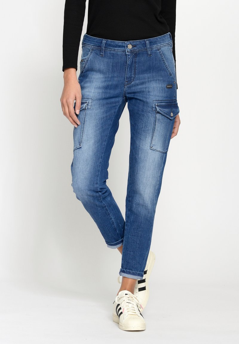 Personne en jean cargo bleu avec grandes poches latérales, haut noir à manches longues et baskets blanches à rayures noires, debout de façon décontractée.