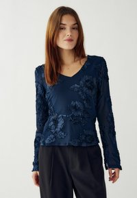 Tumši zila ziedu raksta garām piedurknēm blūze ar V veida kakla izgriezumu, kurai ir struktūrēta auduma ar izcilu izšuvumu un caurspīdīgām piedurknēm, apvienota ar melnām biksēm.