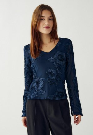 Tamsiai mėlyna gėlių ilgarankovė palaidinė su V formos iškirpte, su tekstūruota medžiaga, iškiliais siuvinėjimais ir permatomomis rankovėmis, derinama su juodais kelnais.