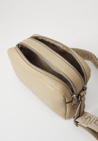 Beige lærveske med glatt tekstur, som har tre glidelåslommer og et stoffbånd med merke.