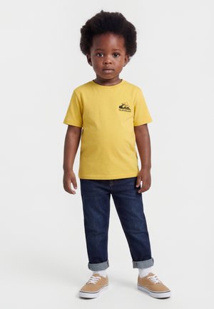 Kisgyermek afro frizurával, sárga rövid ujjú pólót, sötét felhajtott farmert, fehér zoknit és bézs tornacipőt visel, fehér háttér előtt áll.