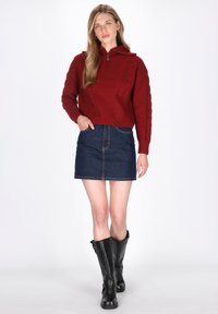 Roter Cable-Knit-Pullover mit kurzem Reißverschluss und langen Ärmeln, kombiniert mit einem dunklen Denim-Rock und knielangen schwarzen Stiefeln.