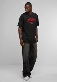 Black cotton t-shirt with red graphic text on the front, paired with loose dark denim jeans and white sneakers. Minimal background.