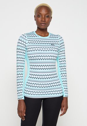 Frau mit kurzen blonden Haaren, die ein eng anliegendes hellblaues Langarmshirt mit schwarzen Zickzackmustern und schwarze Hosen trägt, in neutraler Pose stehend.