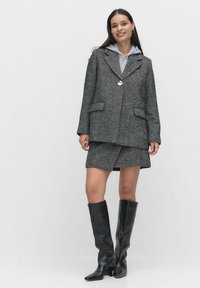 Harmaan kalanruotoblazerin ja hameen asu, jossa on päällä vaalea harmaa huppari. Mallilla on yllään pitkät mustat nahkasaappaat, jotka korostavat rakenteellista muotoilua.