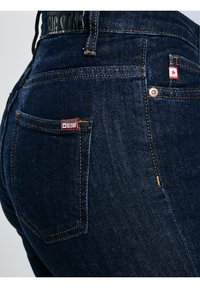 Tuvuņš tumši ziliem džinsa biksēm, kurās redzama aizmugurējā kabata, oranža šuve un sarkans uzlīmes ar baltu zvaigzni un tekstu "BIG STAR".