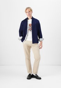 Camisa de botones azul marino sobre una camiseta gráfica blanca, pantalones beiges y mocasines negros. Conjunto casual con forro de camisa a cuadros.