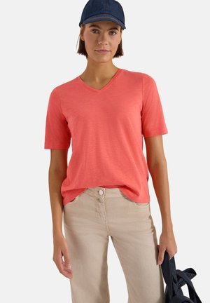 Kobieta w koralowym T-shircie z dekoltem w serek i beżowych spodniach, trzyma granatową kurtkę, na głowie ciemnoniebieska czapka z daszkiem na białym tle.