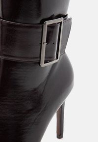 Gros plan d'une botte en cuir noir à talon haut avec une large sangle et une boucle rectangulaire argentée autour de la cheville.
