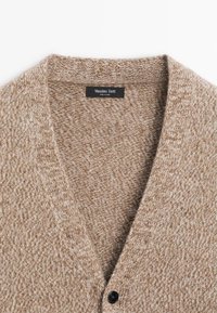 Cardigan lavorato a maglia beige con scollatura a V, caratterizzato da un motivo testurizzato e un singolo bottone nero. L'etichetta reca la scritta "Massimo Dutti."