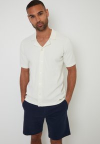 Polo shirt blanc à manches courtes en tricot avec un col et une patte de boutonnage, associé à un short marine. Tissu texturé avec un motif subtil.