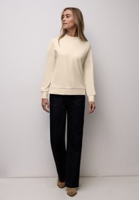 Sweat-shirt crème à col rond avec épaules tombantes, associé à un jean large foncé. Des bottines beige complètent la tenue.