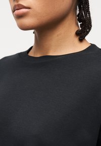 Άτομο με κοτσίδες που φοράει μια απλή μαύρη μπλούζα με στρογγυλή λαιμόκοψη, εμφανίζοντας το κάτω μέρος του προσώπου, τον λαιμό και το επάνω μέρος του στήθους.