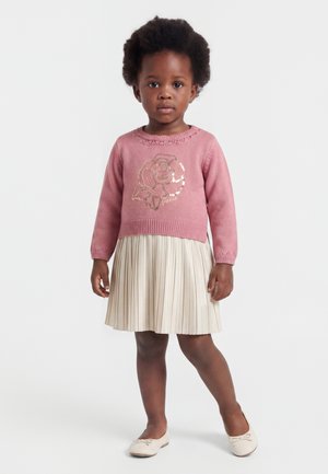 Bambino piccolo con capelli ricci che indossa un maglione rosa con un motivo di rosa, gonna beige plissettata e ballerine crema, in piedi su sfondo bianco.