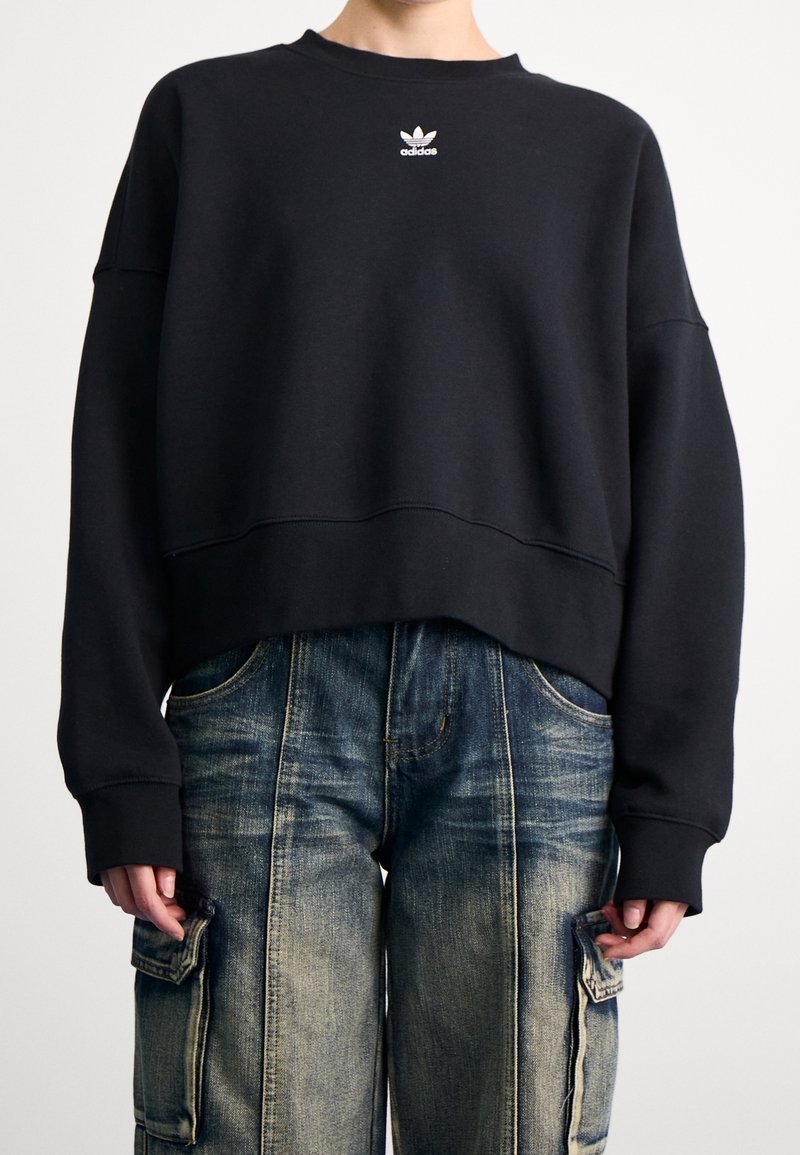 Schwarzes, verkürztes Sweatshirt mit rundem Ausschnitt und langen Ärmeln, ausgestattet mit dem weißen Adidas-Logo auf der Brust, kombiniert mit hellblauen Denim-Cargohosen.