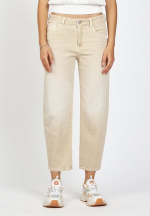 Woman wearing beige straight-leg jeans, white crop top, and white sneakers with orange accents, standing against plain background.