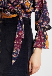 Multicolor bloemenblouse met lange mouwen en een strikzoom, gecombineerd met een donkere corduroy rok met knopen aan de voorkant en een geribbelde textuur.
