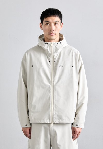 Chaqueta con capucha en un beige claro, con cierre de cremallera y confeccionada en un tejido suave. Presenta detalles en negro y un diseño de corte ligeramente holgado.