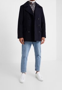Parker in pura lana navy con design doppiopetto, grandi bottoni e tasche laterali. Abbinato a jeans azzurri chiari e sneakers bianche.